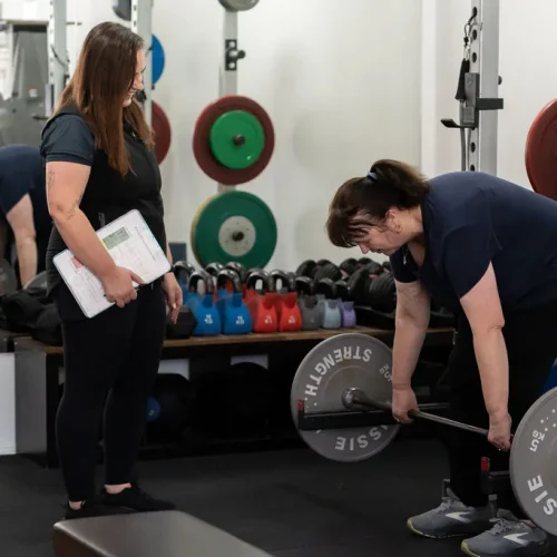 Exercise physiologist assisting as woman completes deadlift with barbell in a squat rack