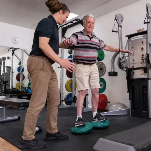 An older gentleman is balancing while standing on green wobble pads under his feet, with an Exercise Physiologist Standing next to him to support if needed